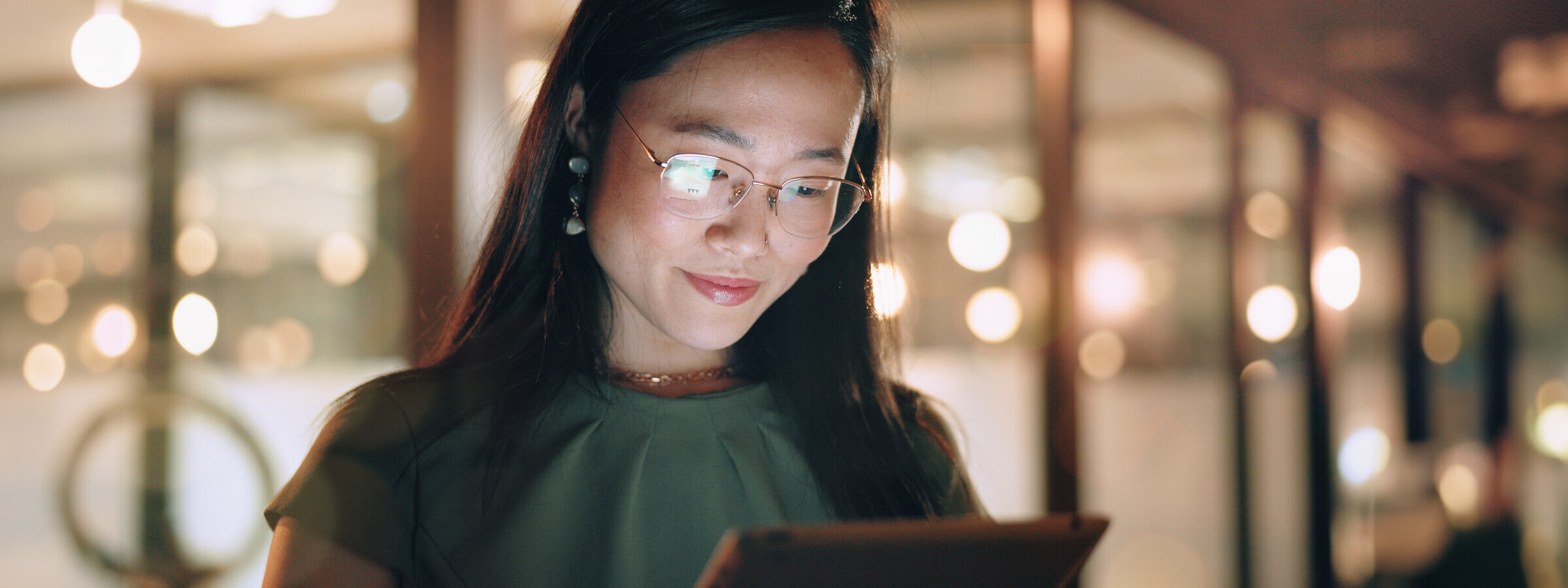 Eine Frau mit Brille schaut l&auml;chelnd in ein Tablet. Im Hintergrund sind beleuchtete Fenster eines Geb&auml;udes zu erkennen. 