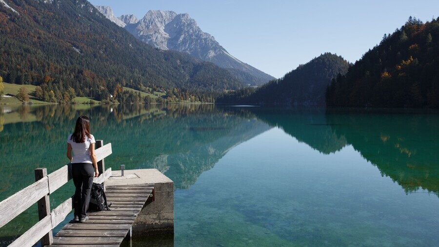 Ein Holzsteg reicht in einen blauen Gebirgssee, der umgeben von Bergen und Wäldern ist. 