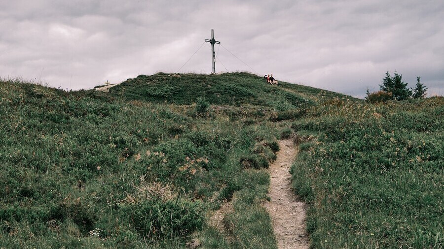 Schmaler Wanderweg führt über eine alpine Wiese zu einem Gipfelkreuz.