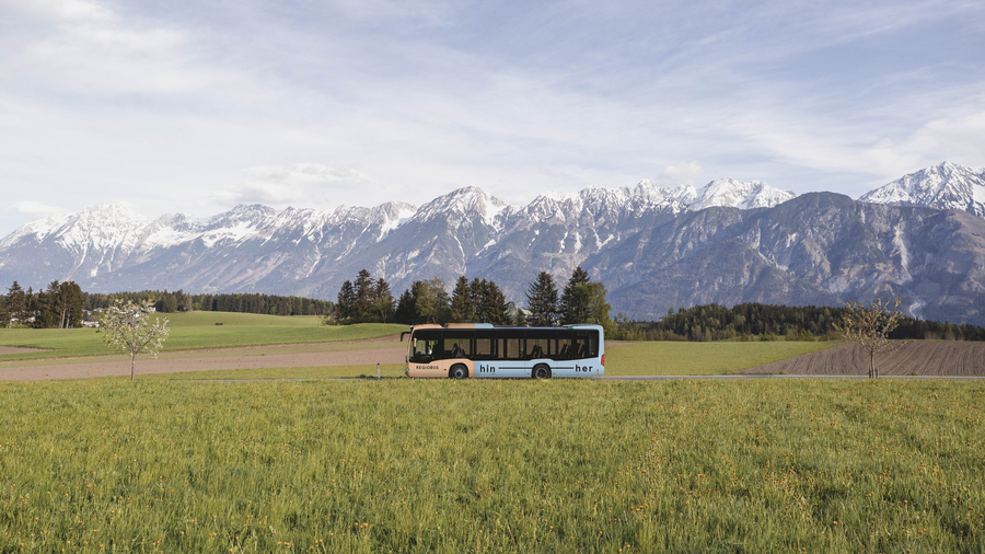 Ein orange-blauer Bus fährt auf einer grünen Wiese. im Hintergrund sieht man eine Bergkette mit Schnee auf den Spitzen.