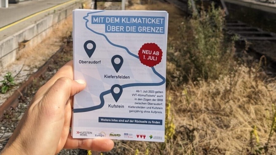 Ein Flyer mit dem folgenden Informationen wird in der Hand gehalten: "Mit dem Klimaticket über die Grenze. Ab 1. Juli 2023 gelten VVT-KlimaTickets auch in den Zügen der BRB zwischen Oberaudorf, Kiefersfelden und Kufstein ganzjährig ohne Aufpreis.".